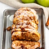 A sliced loaf of apple cinnamon bread drizzled with white icing sits on a metal tray. In the background, there are three green apples and a cinnamon stick on a beige cloth.