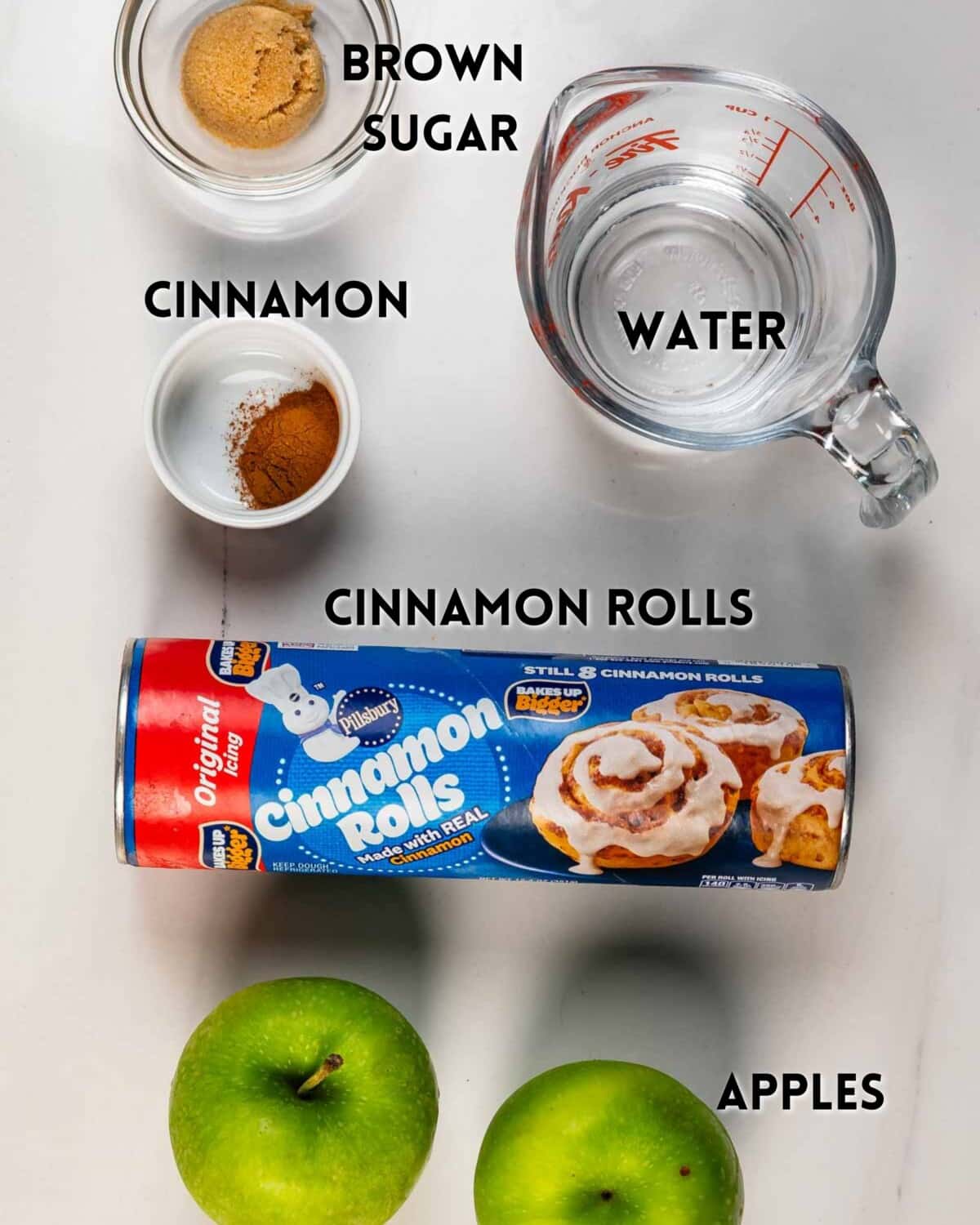 Flat lay of ingredients on a white surface: a bowl of brown sugar, a bowl of cinnamon, a measuring cup of water, a can of cinnamon rolls, and two green apples, each labeled with text.