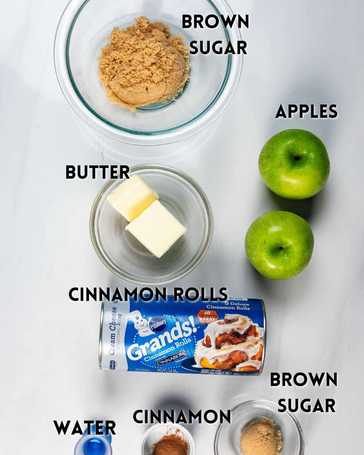 Ingredients for a recipe arranged on a white surface: a bowl of brown sugar, two green apples, butter in a small dish, a can of Grands! cinnamon rolls, water, ground cinnamon, and another bowl of brown sugar.