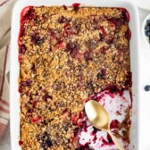 A baked mixed berry crisp in a white rectangular dish with a golden crumbly topping. A serving has been scooped out, showing the juicy berry filling and a gold spoon rests inside the dish. Nearby are a towel and a few loose berries.