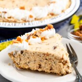 A slice of creamy pie on a white plate, topped with whipped cream and crumbled bits. The pie appears to contain nuts or chocolate chips. The plate is placed beside a fork, with the rest of the pie visible in the background.