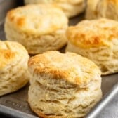 A baking tray with freshly baked golden-brown biscuits, showcasing their flaky, layered texture. The biscuits are arranged closely together on a silver tray, on a gray surface.