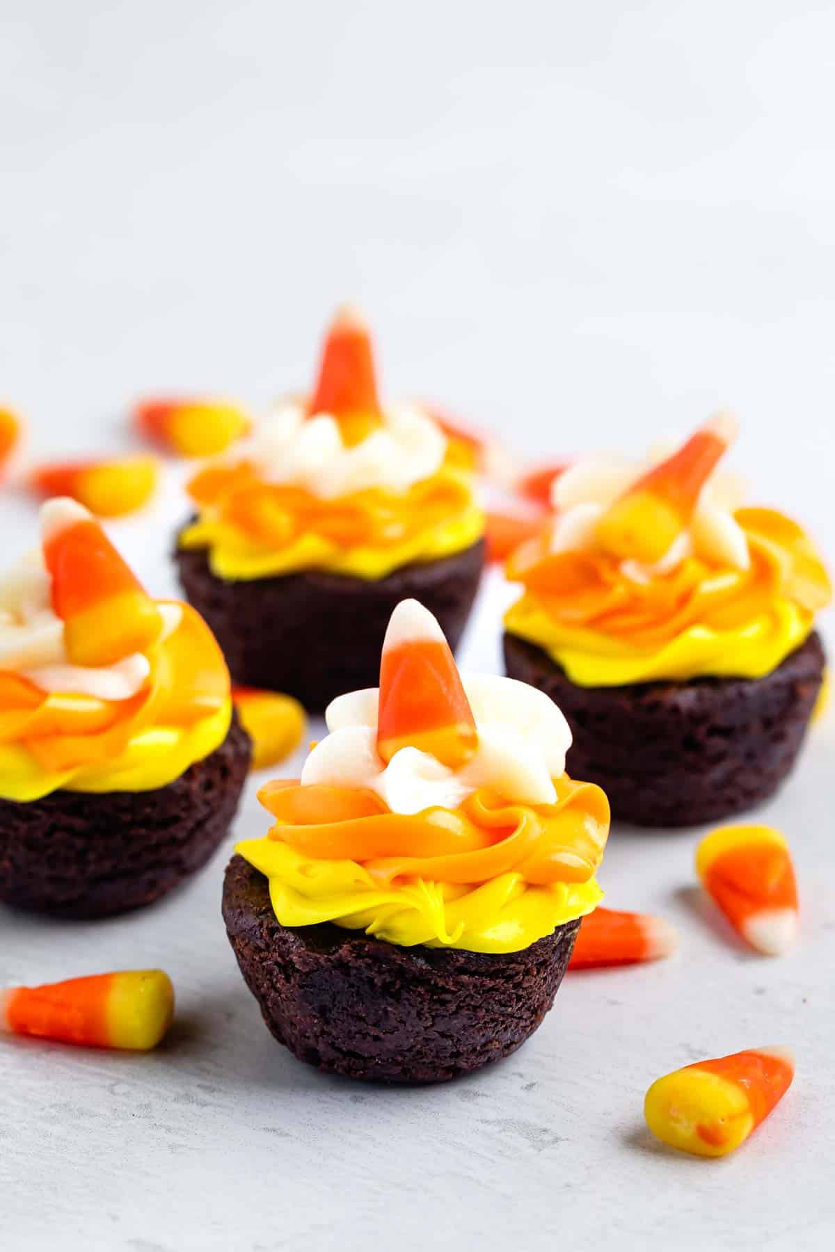 Several chocolate brownie bites topped with yellow and orange icing, a swirl of white frosting, and a piece of candy corn.