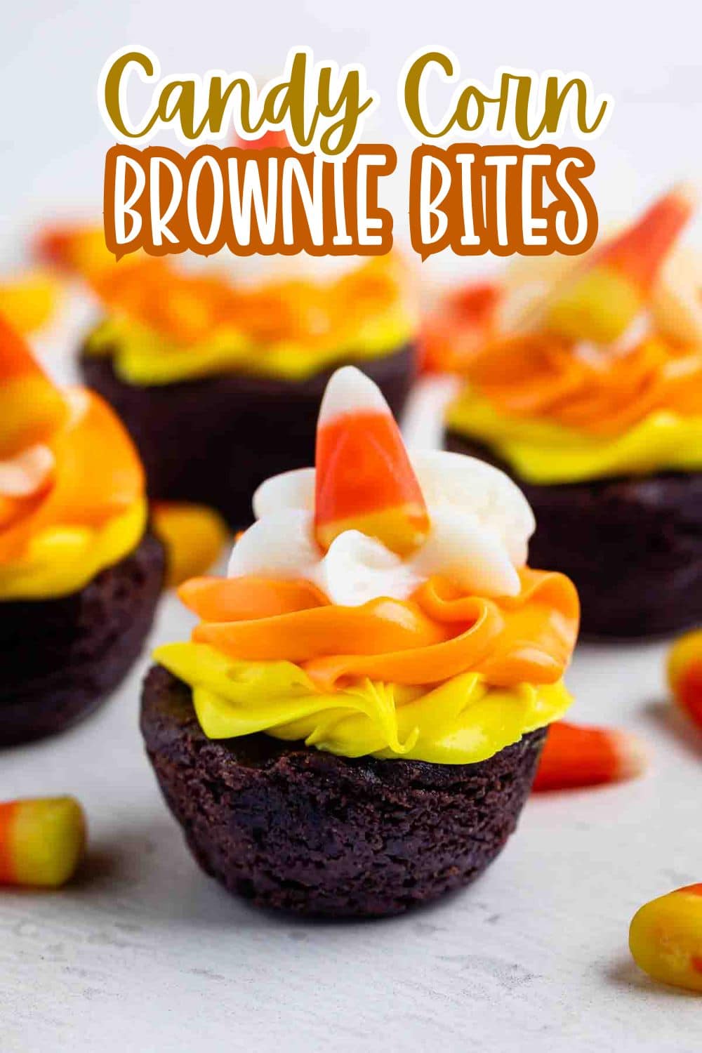Several chocolate brownie bites topped with yellow and orange icing, a swirl of white frosting, and a piece of candy corn. Text at the top reads “Candy Corn Brownie Bites.”.