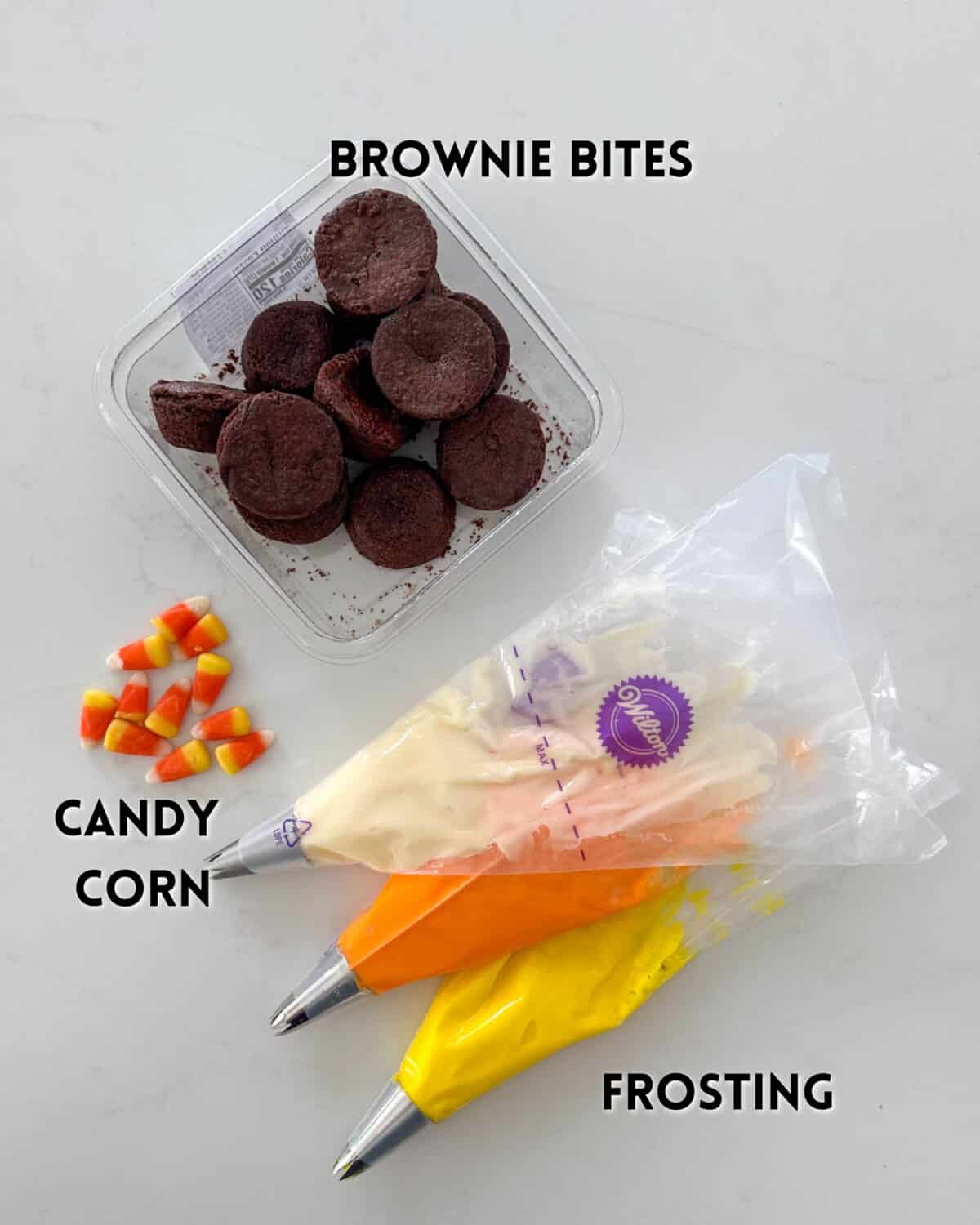 A container of brownie bites, candy corn pieces, and three piping bags filled with white, orange, and yellow frosting on a white surface. Labels identify each item: BROWNIE BITES, CANDY CORN, and FROSTING.