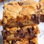 A close-up of three stacked chocolate chip cookie bars filled with gooey caramel and melted chocolate chips, with a soft and crumbly texture. Unwrapped caramel candies are visible in the background.