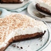 A chocolate cream pie with a slice removed, revealing layers of creamy chocolate filling topped with whipped cream and chocolate shavings. Two plates in the background hold slices of the pie with forks beside them.