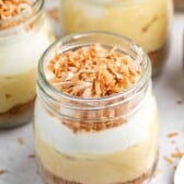 Glass jars filled with layers of creamy pudding, topped with a dollop of whipped cream and sprinkled with toasted coconut flakes. The dessert sits on a light surface, with a spoon nearby.
