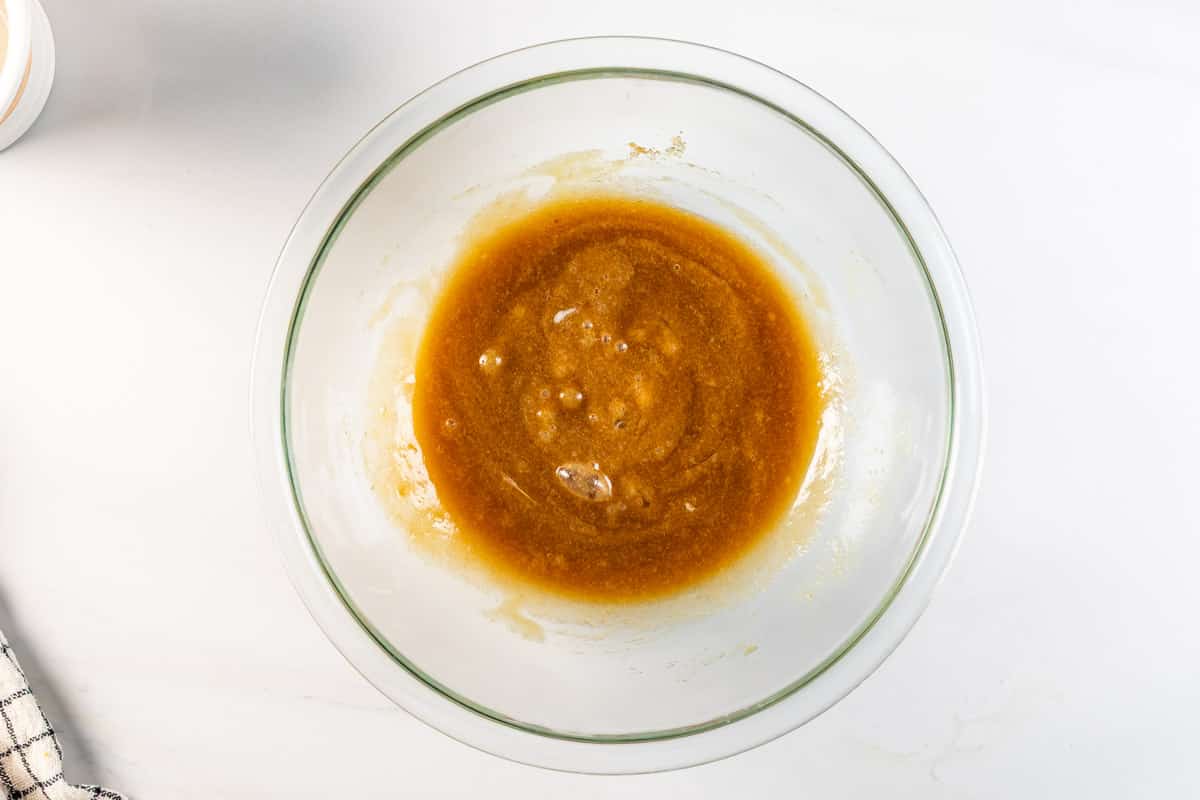 A glass mixing bowl containing a thick, caramel-colored mixture sits on a white surface, with a checkered kitchen towel partially visible in the bottom left corner.