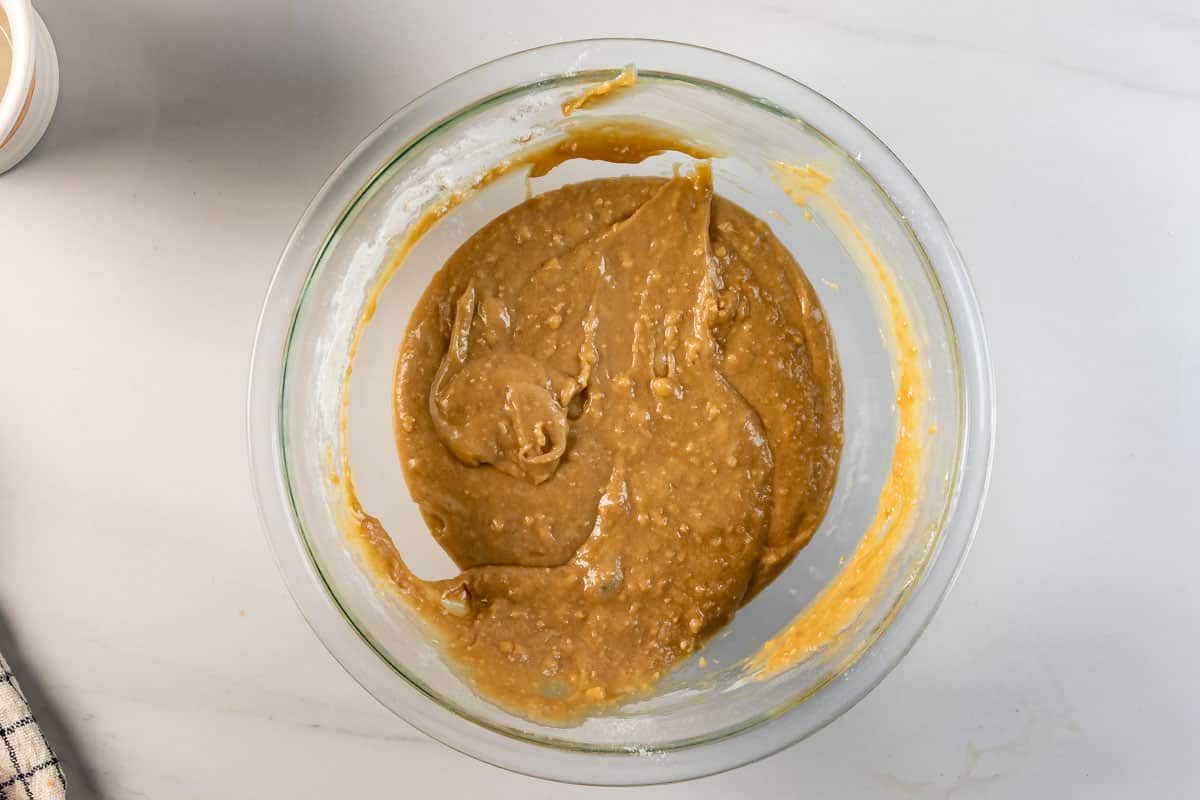 A glass bowl filled with thick, light brown banana bread batter sits on a white countertop; a bit of batter clings to the sides of the bowl.