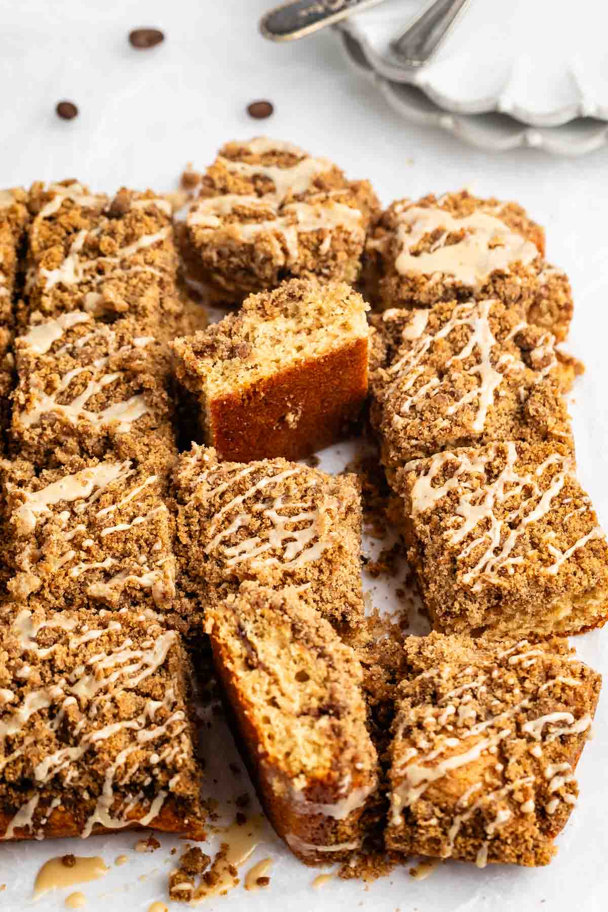 Rectangular slices of coffee cake topped with brown crumbly streusel and white icing drizzle, arranged on a white surface. Some slices are stacked, showing the moist, dense texture inside.