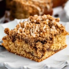 A slice of crumb cake with a thick layer of cinnamon streusel topping and a light drizzle of icing, served on a white decorative plate.