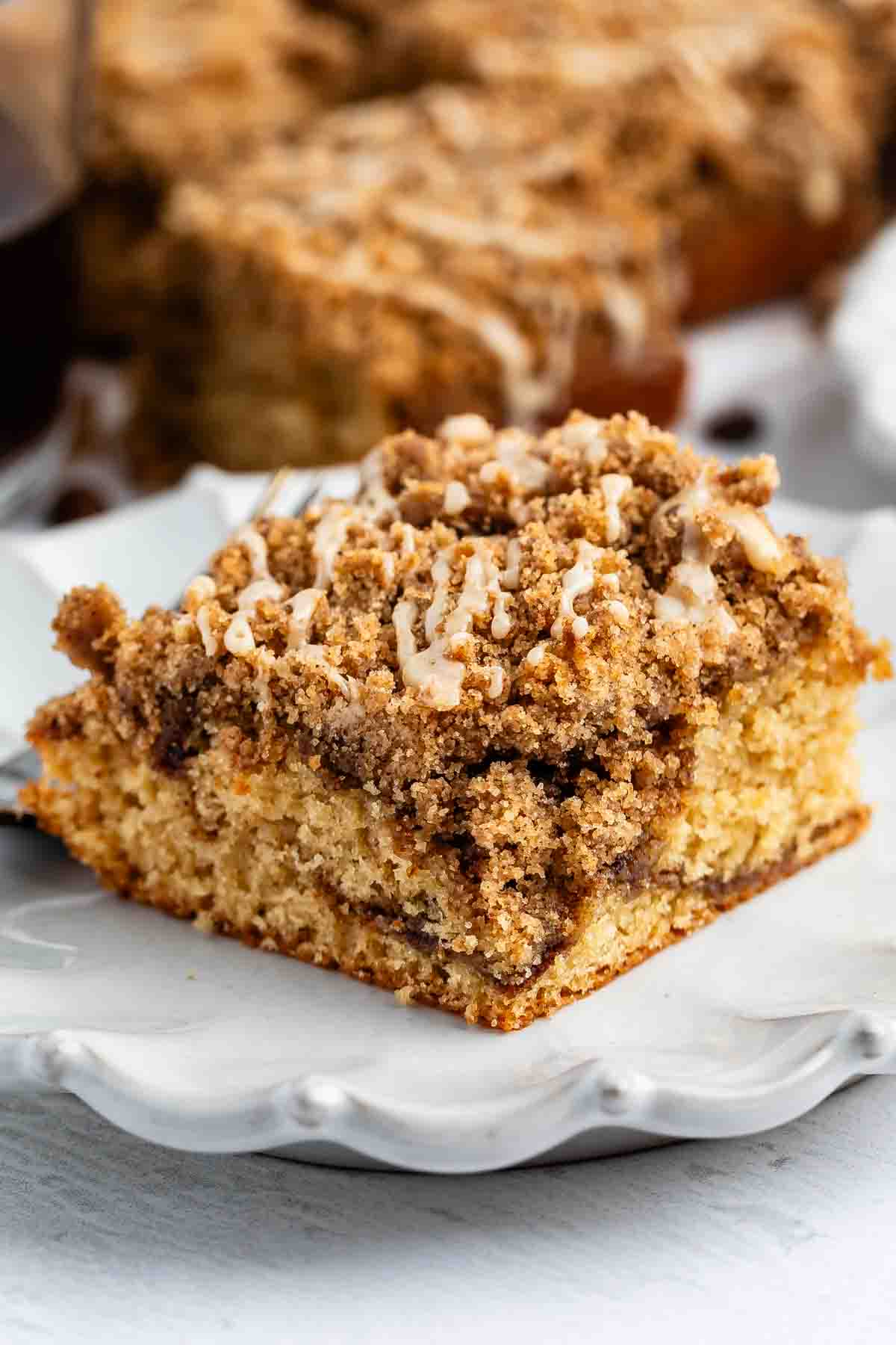 A slice of crumb cake with a thick layer of cinnamon streusel topping and a light drizzle of icing, served on a white decorative plate.
