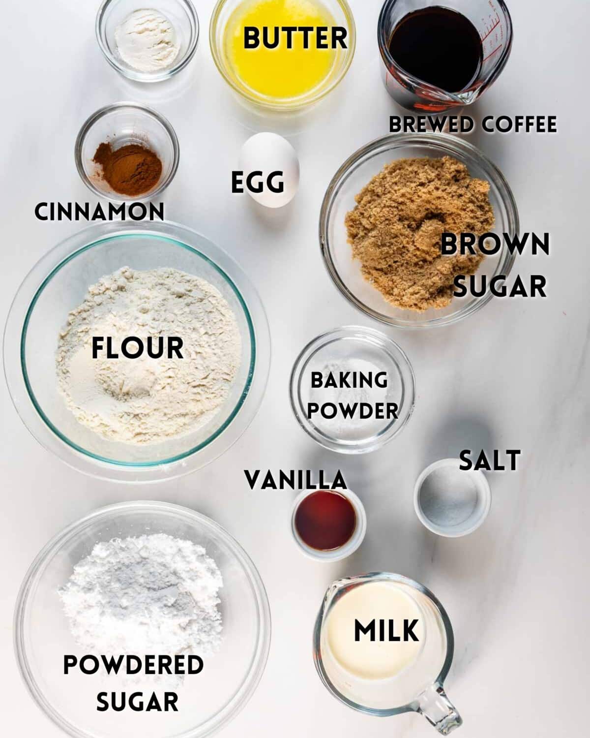 Top-down view of baking ingredients in bowls: butter, brewed coffee, brown sugar, flour, cinnamon, baking powder, egg, salt, vanilla, milk, and powdered sugar, all labeled on a white surface.