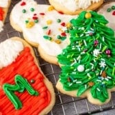 Cut Out Sugar Cookies on a cooling rack, decorated with colorful icing: a red stocking with a green M, a green Christmas tree with sprinkles, and a white cookie topped with multicolored sprinkles.