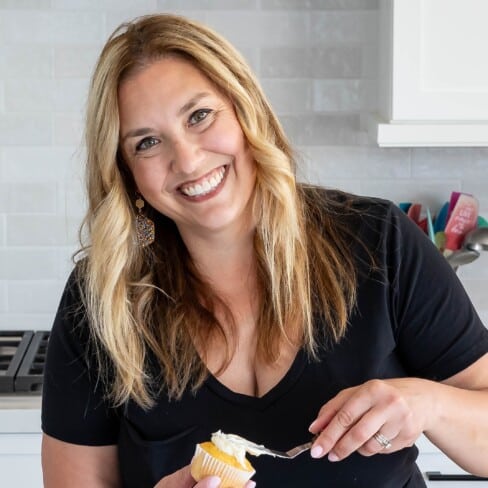 A smiling woman in a black shirt is all about perfecting her cupcake, spreading frosting with care in a modern kitchen. White cabinets and a sleek stovetop provide the perfect backdrop for her culinary creation.