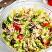 A glass bowl filled with Greek or Mediterranean salad containing lettuce, cherry tomatoes, cucumber, red onion, chickpeas, black olives, feta cheese, and orzo, with wooden utensils and tomatoes in the background.