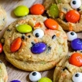 Colorful Halloween cookies topped with orange, yellow, green, and purple candy-coated chocolates and candy eyeballs are arranged on a wooden surface, with some loose candies and candy eyes scattered around the cookies.
