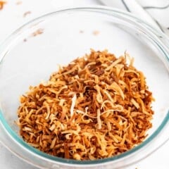A clear glass bowl filled with golden-brown toasted shredded coconut sits on a white surface, with a striped cloth in the background and a few coconut flakes scattered nearby.