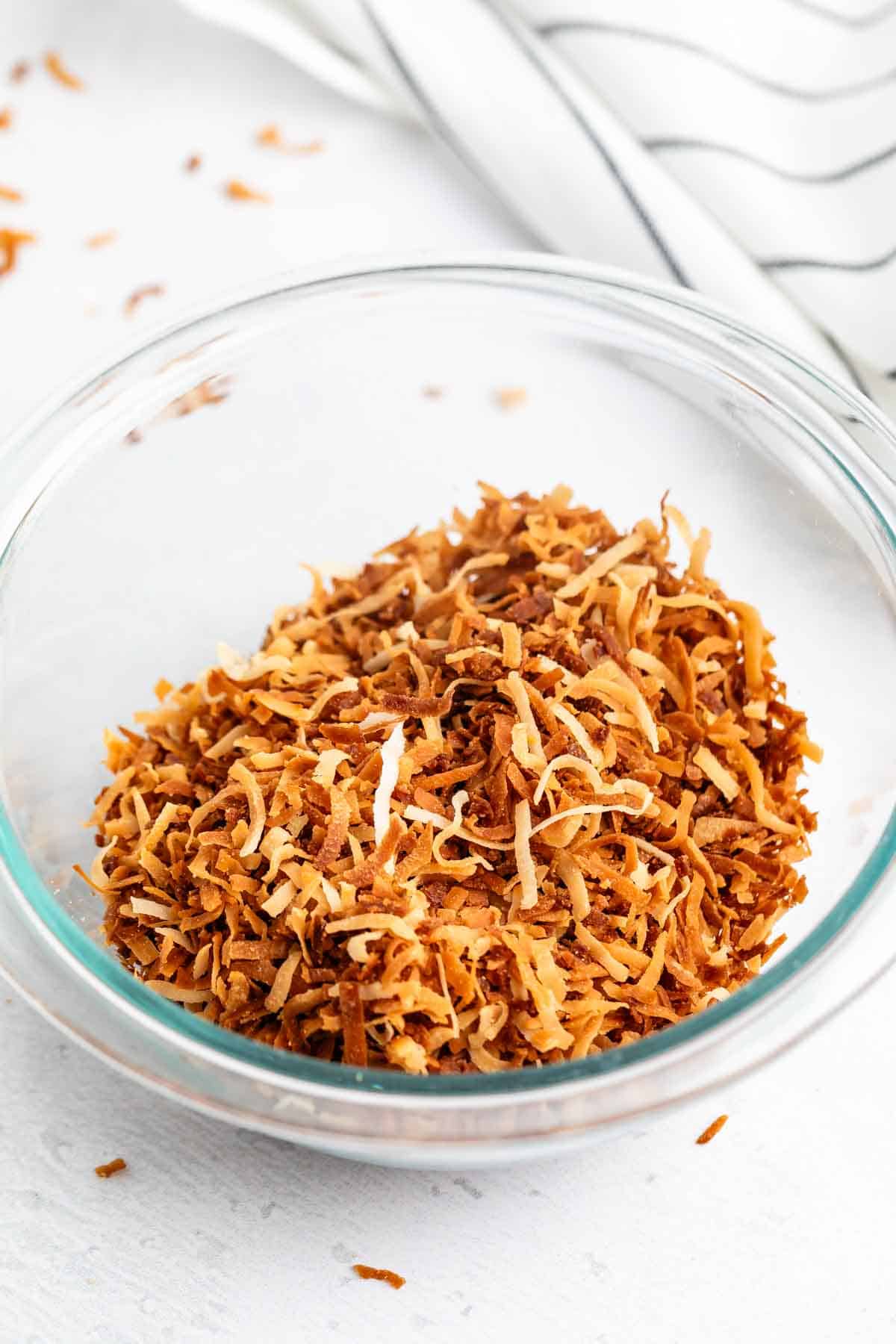 A clear glass bowl filled with golden-brown toasted shredded coconut sits on a white surface, with a striped cloth in the background and a few coconut flakes scattered nearby.