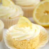 A lemon cupcake with creamy white frosting and a small lemon wedge on top, sitting on an unwrapped cupcake liner. Other cupcakes and a halved lemon are visible in the background.