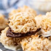Close-up of several macaroon cookies coated with dark and white chocolate on the bottom, resting on a cooling rack. The toasted coconut gives them a golden-brown appearance. A blurred background suggests more macaroons.