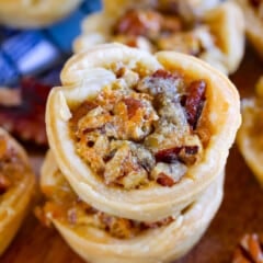 Close-up of stacked mini pecan pies on a wooden surface, showing flaky pastry filled with caramelized pecans. Out-of-focus tarts and a cooling rack appear in the background.