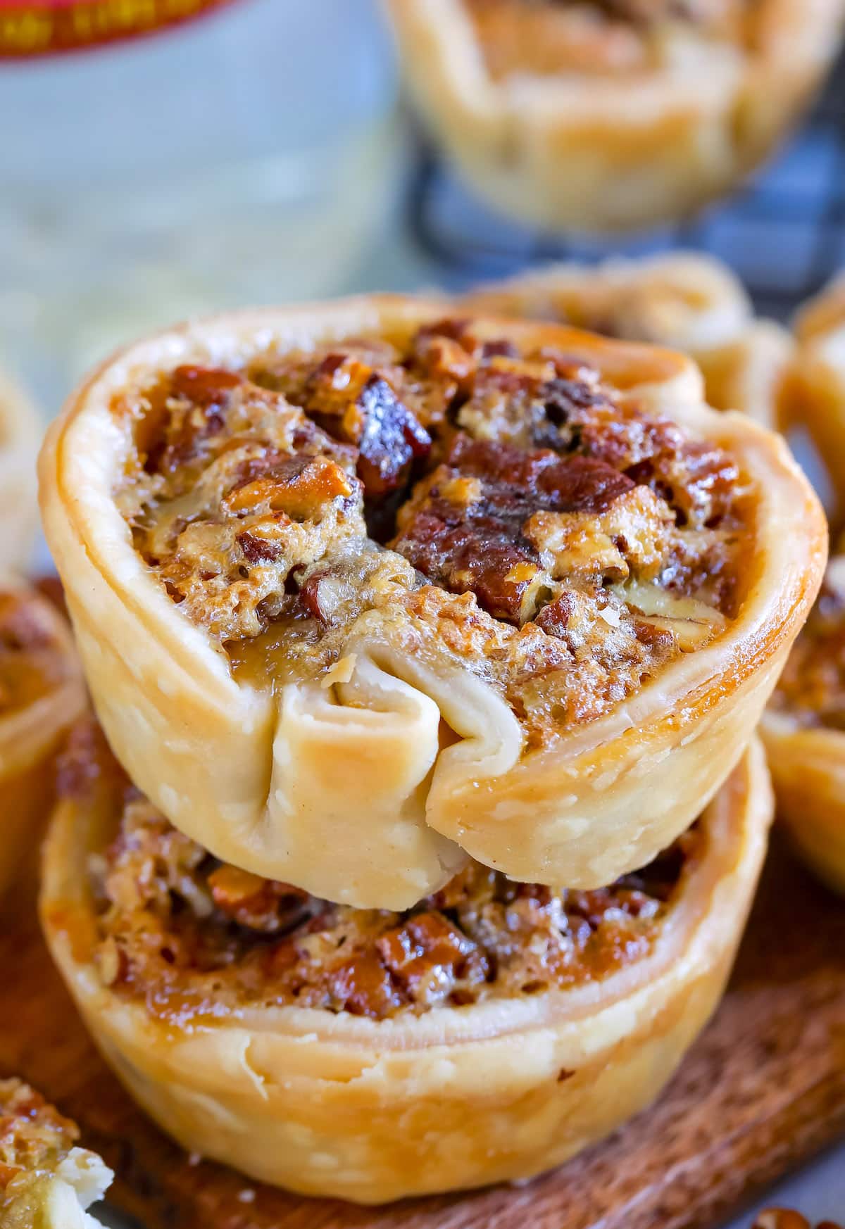 Two mini pecan pies with golden, flaky crusts are stacked on a wooden surface. Their glossy, caramelized filling is studded with chopped pecans. More mini pecan pies are blurred in the background, creating a cozy, inviting scene.