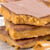 Three peanut butter bars with a chocolate topping are stacked on parchment paper. A spoonful of creamy peanut butter sits in the foreground. The bars have a thick, crumbly base and smooth chocolate layer.
