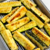 A baking tray filled with golden-brown, breaded zucchini sticks arranged in rows, showing a crispy texture and seasoning on the surface.