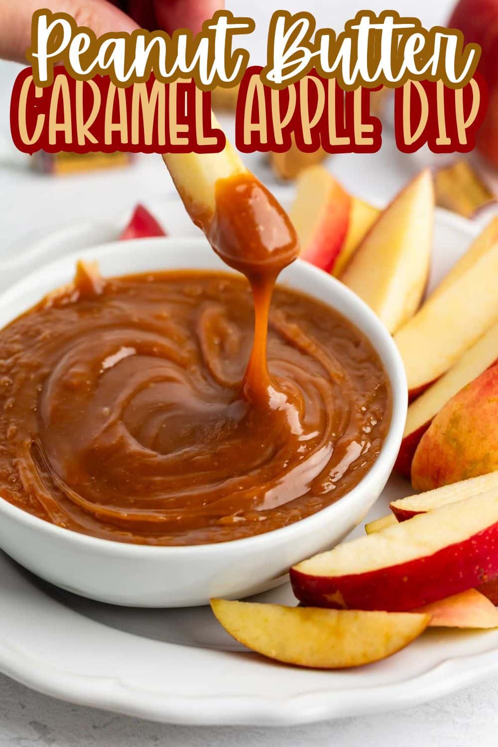 A hand dips an apple slice into a bowl of creamy peanut butter caramel dip, with more apple slices arranged on a white plate. The text Peanut Butter Caramel Apple Dip is displayed at the top.