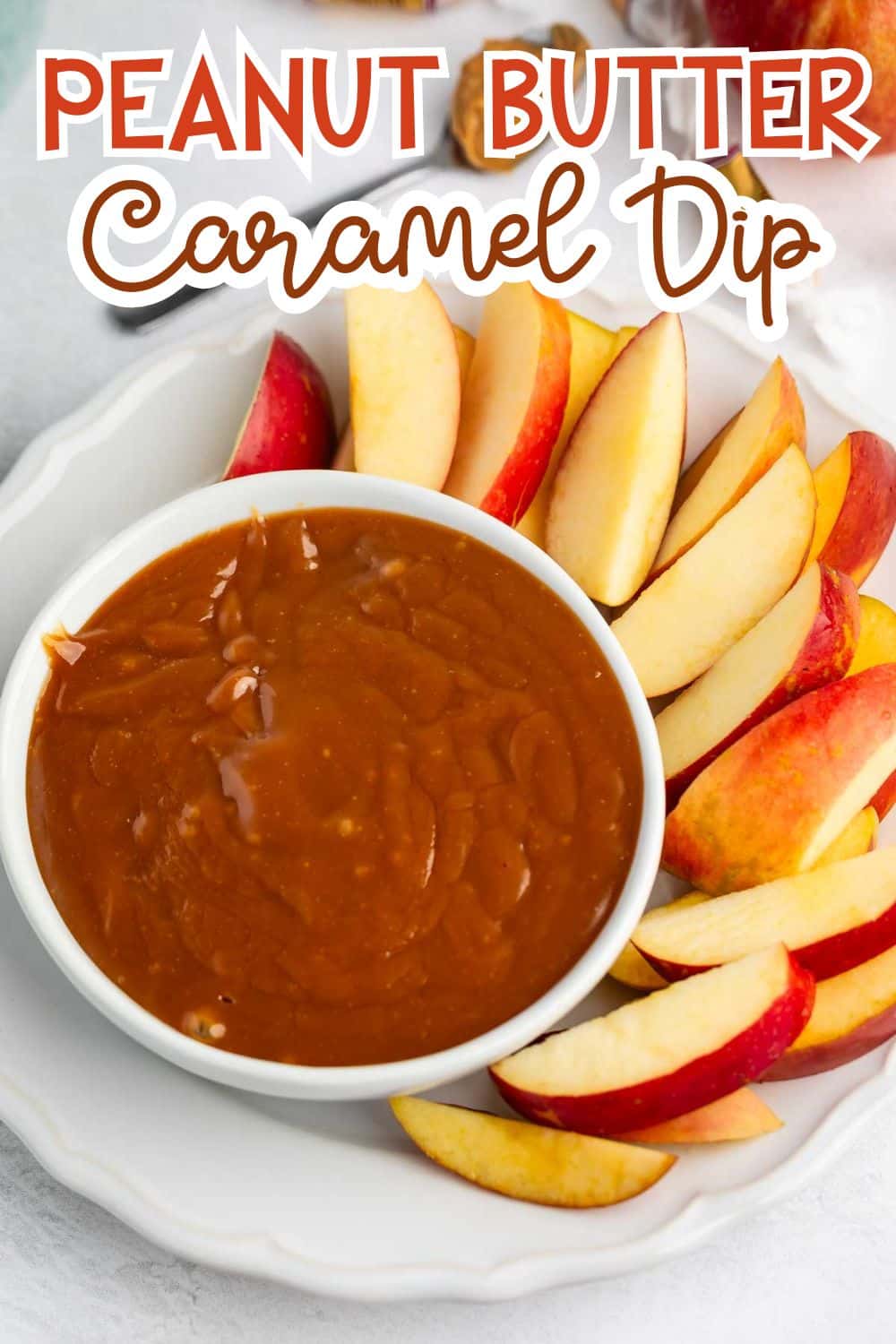 A white plate holds a bowl of peanut butter caramel dip surrounded by fresh red apple slices. The text at the top reads “Peanut Butter Caramel Dip.”.