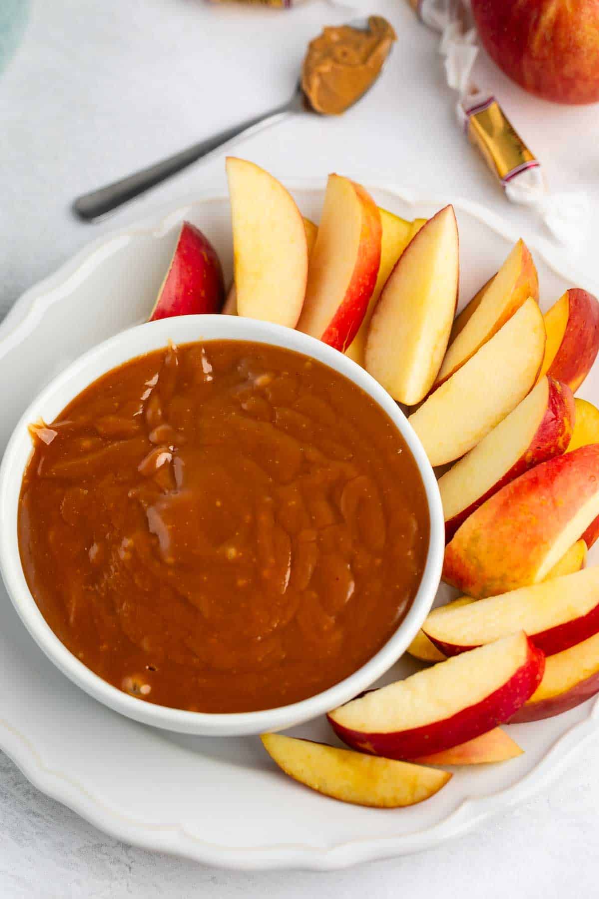 Sliced red apples arranged around a bowl of caramel dip on a white plate, with a spoon and part of an apple visible in the background.