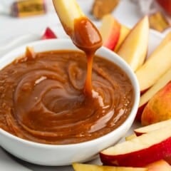 A hand dips a fresh apple slice into a bowl of creamy caramel sauce, surrounded by more apple slices on a white plate. Wrapped caramel candies are visible in the background.