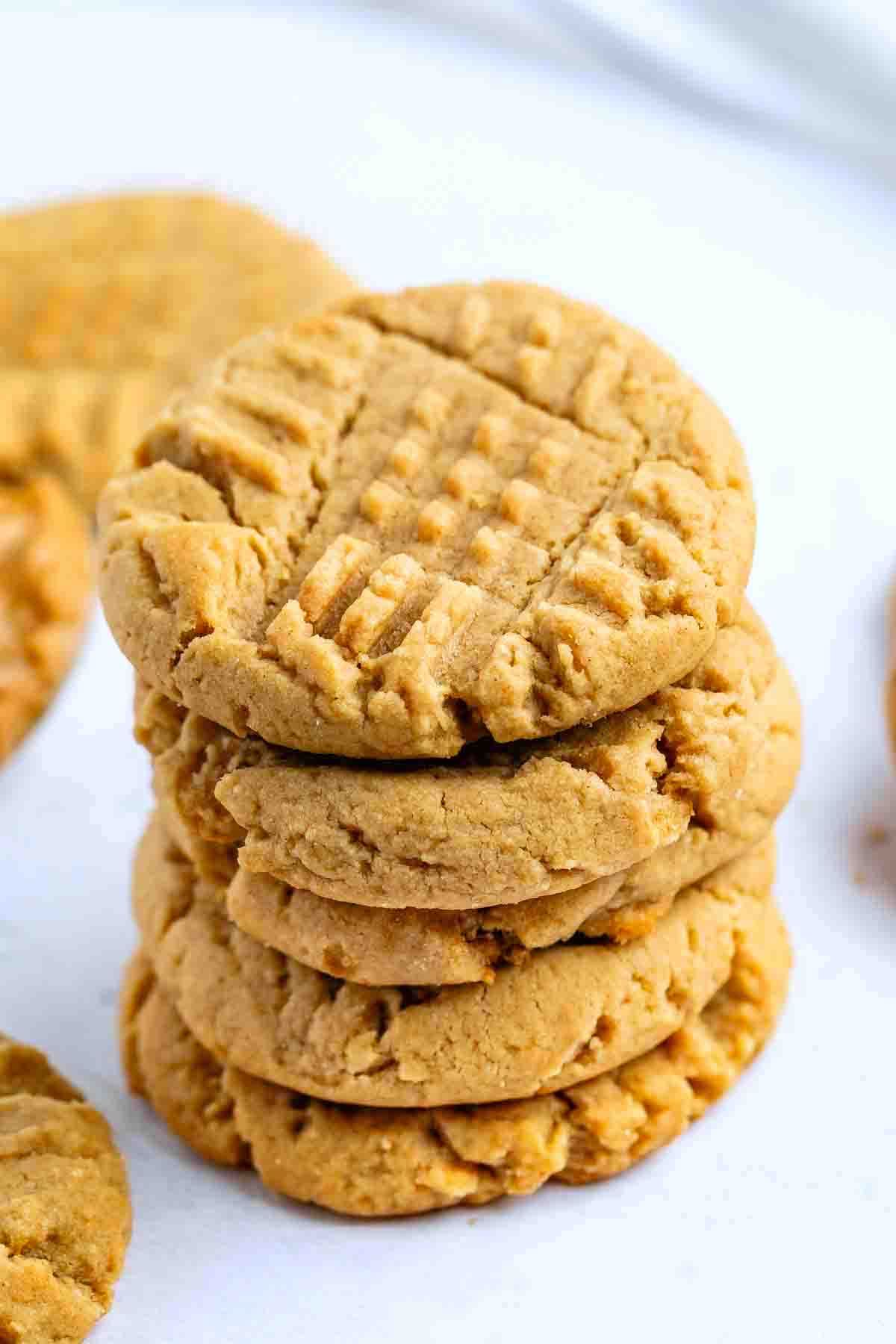 A stack of four golden brown peanut butter cookies with a crisscross fork pattern on top sits on a white surface, tempting anyone searching for the perfect peanut butter cookies recipe. More cookies are blurred in the background.