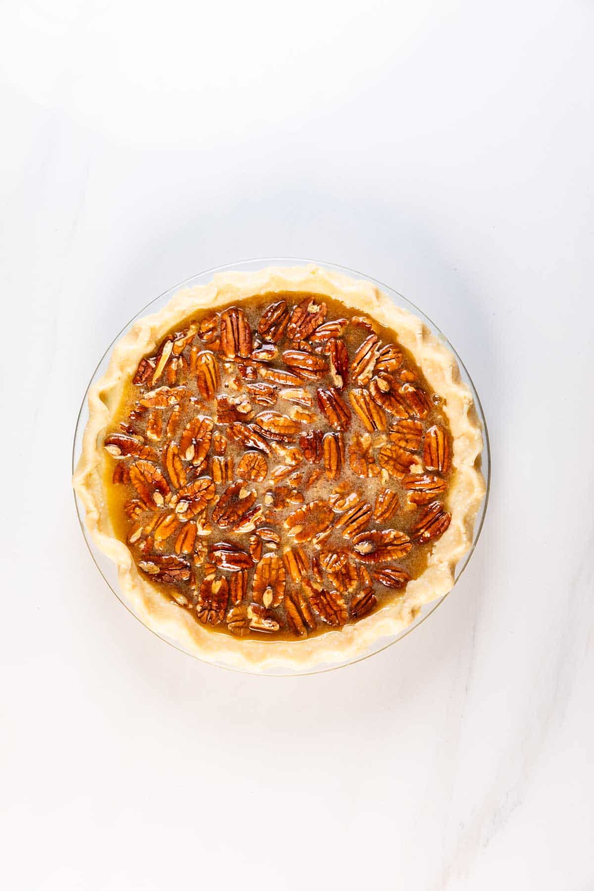 A whole pecan pie with a golden, flaky crust and a glossy filling topped with pecan halves, set on a white marble surface.