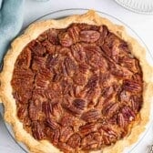 A whole pecan pie with a golden, flaky crust sits on a white surface next to a blue cloth and a white plate. The pie is topped with whole pecan halves arranged over a caramelized filling.