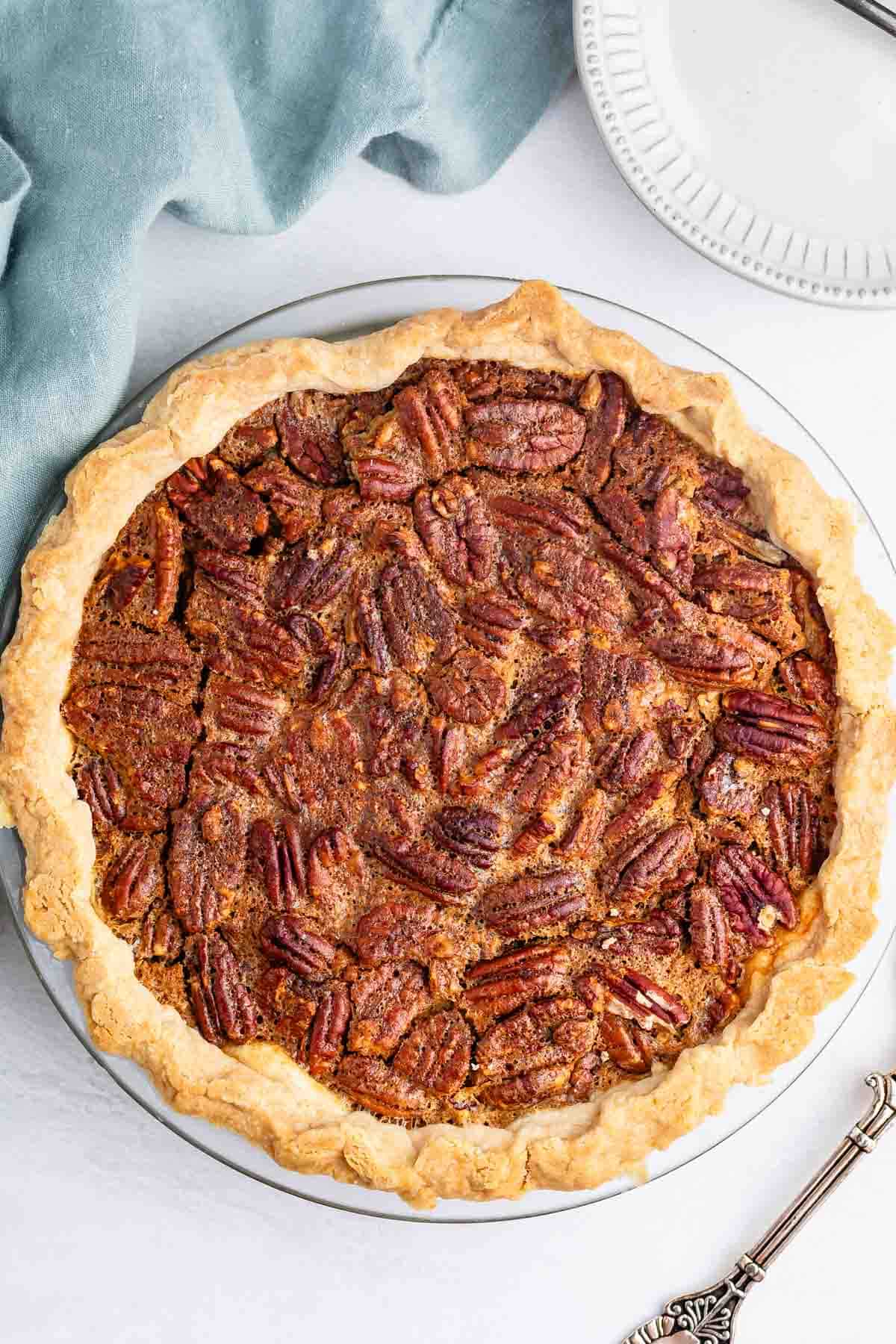 A whole pecan pie with a golden, flaky crust sits on a white surface next to a blue cloth and a white plate. The pie is topped with whole pecan halves arranged over a caramelized filling.