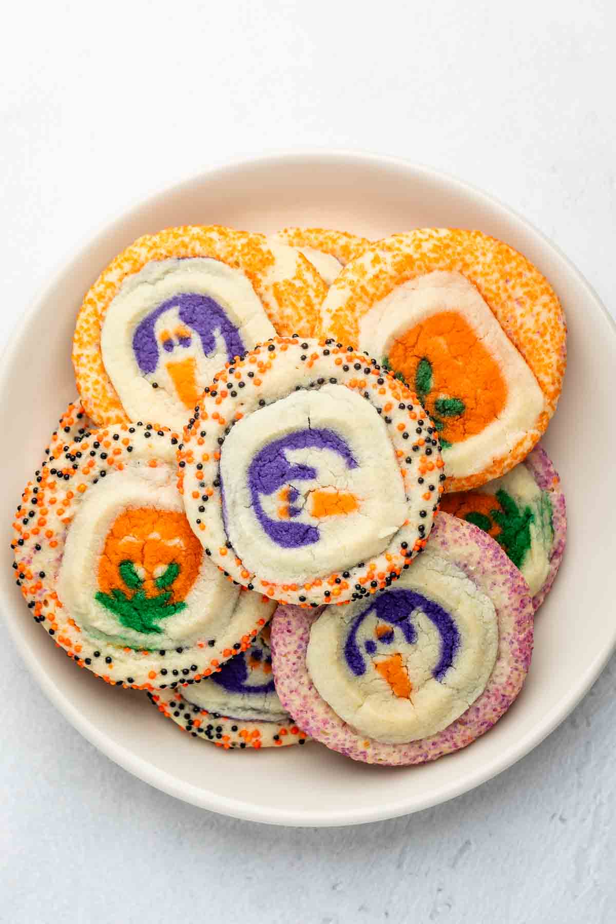 A white bowl filled with colorful Halloween-themed sugar cookies decorated with designs like pumpkins and ghosts, edged with orange, purple, and black sprinkles.