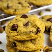 A stack of three chocolate chip cookies with visible chunks of chocolate and pistachios, surrounded by more cookies and scattered pistachios on a white surface. A cooling rack with more cookies is in the background.