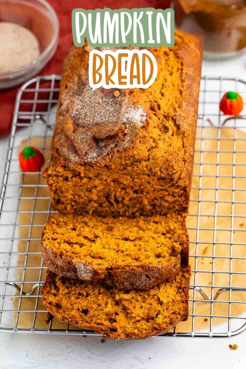 A loaf of pumpkin bread with a sugar topping sits on a wire cooling rack, with two slices cut. Decorative candy pumpkins are nearby. The text Pumpkin Bread is written above the loaf.
