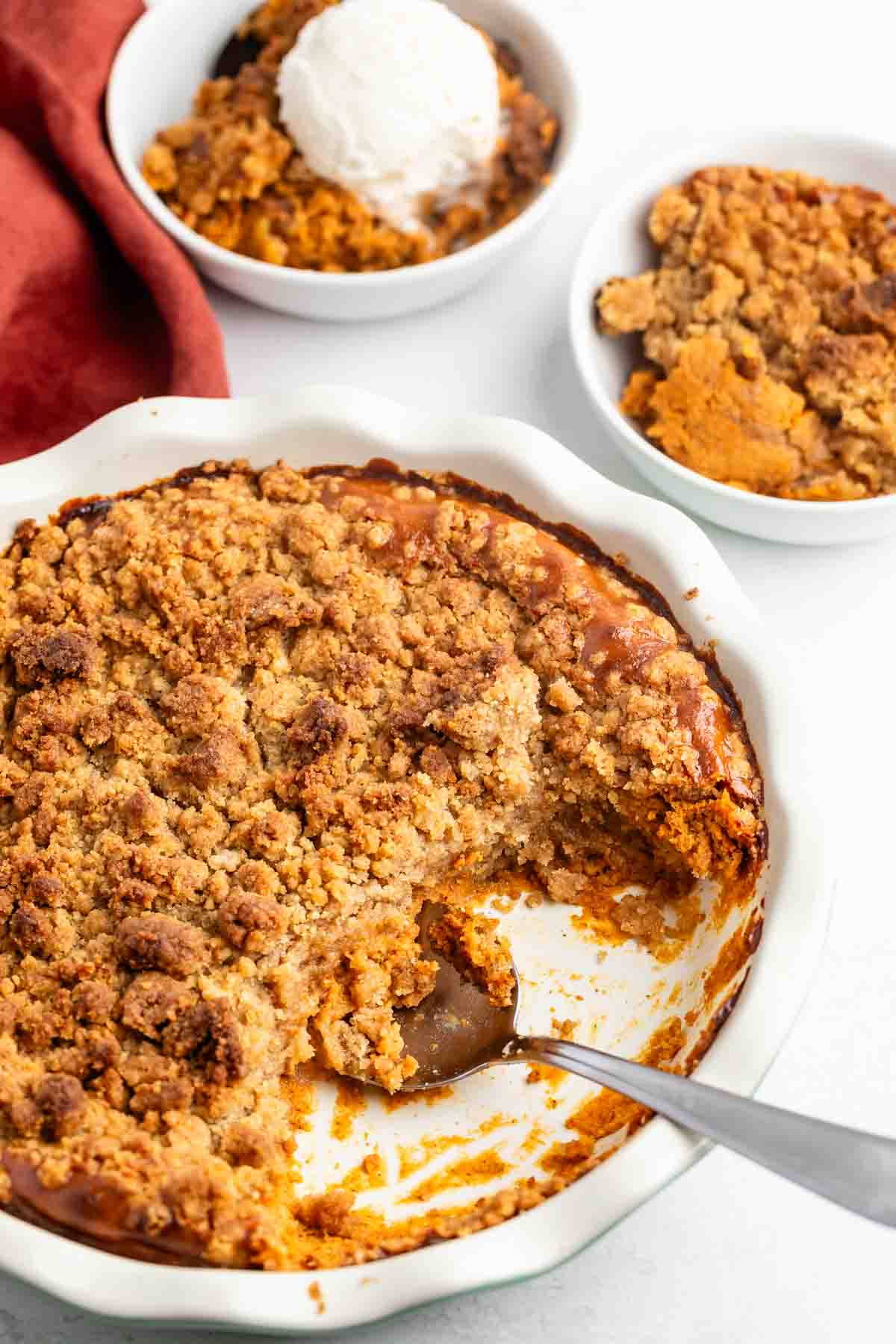 A round baking dish filled with pumpkin crisp topped with a golden crumbly streusel, a large serving scooped out. Nearby are two small bowls of the dessert, one with a scoop of vanilla ice cream.
