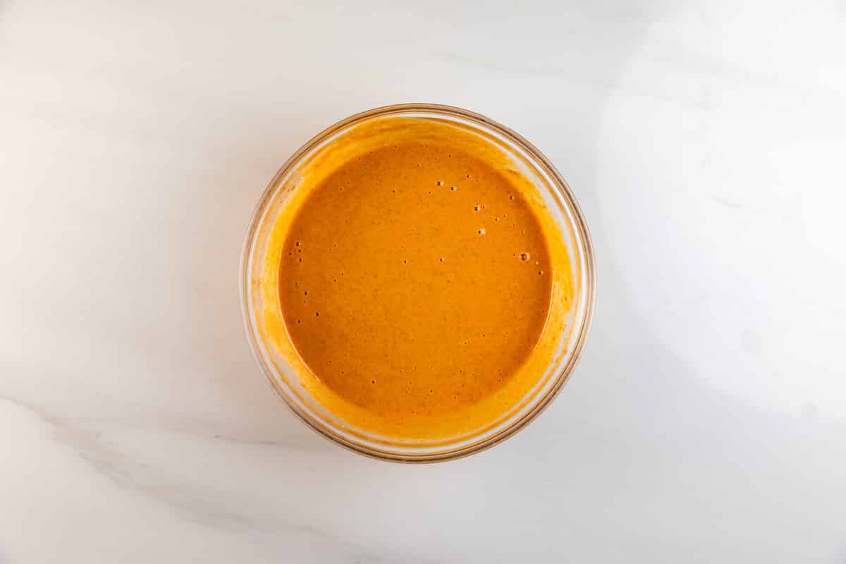 A clear glass bowl filled with smooth, orange-colored sauce sits on a white marble surface.