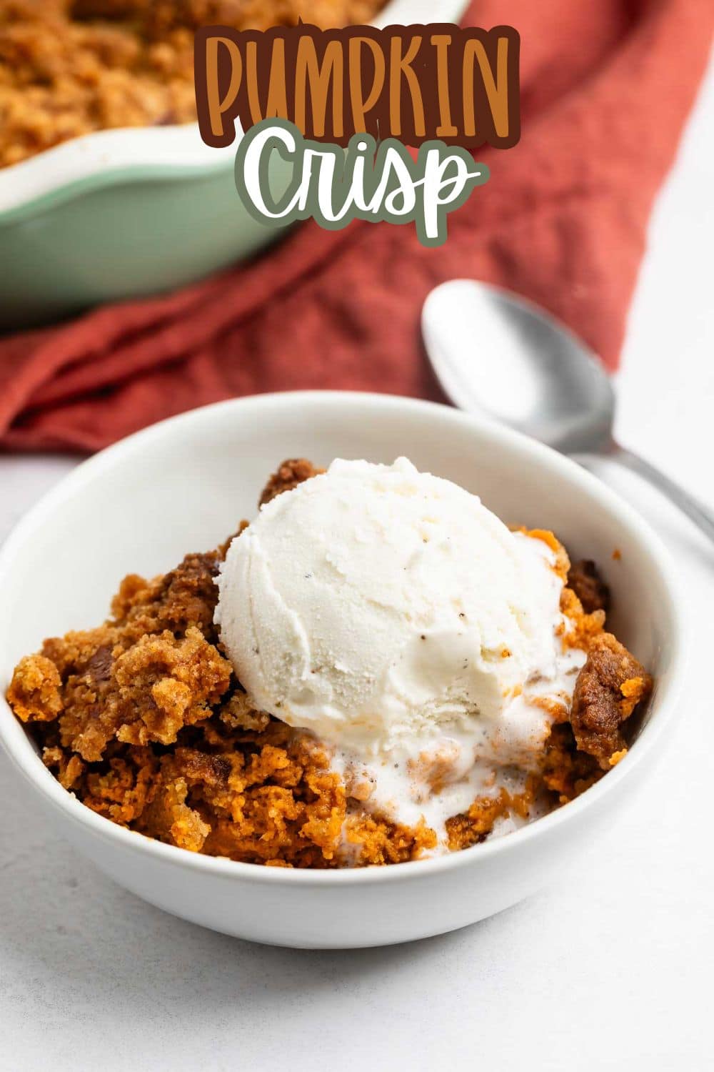 A bowl of pumpkin crisp topped with a scoop of vanilla ice cream, with a spoon beside it. An orange cloth and a baking dish are in the background. Text at the top reads “Pumpkin Crisp.”.