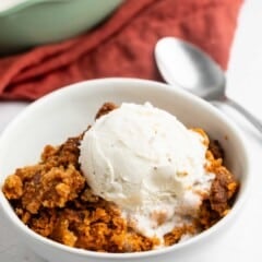 A bowl of pumpkin dump cake topped with a scoop of vanilla ice cream, with a spoon and a red cloth napkin in the background.