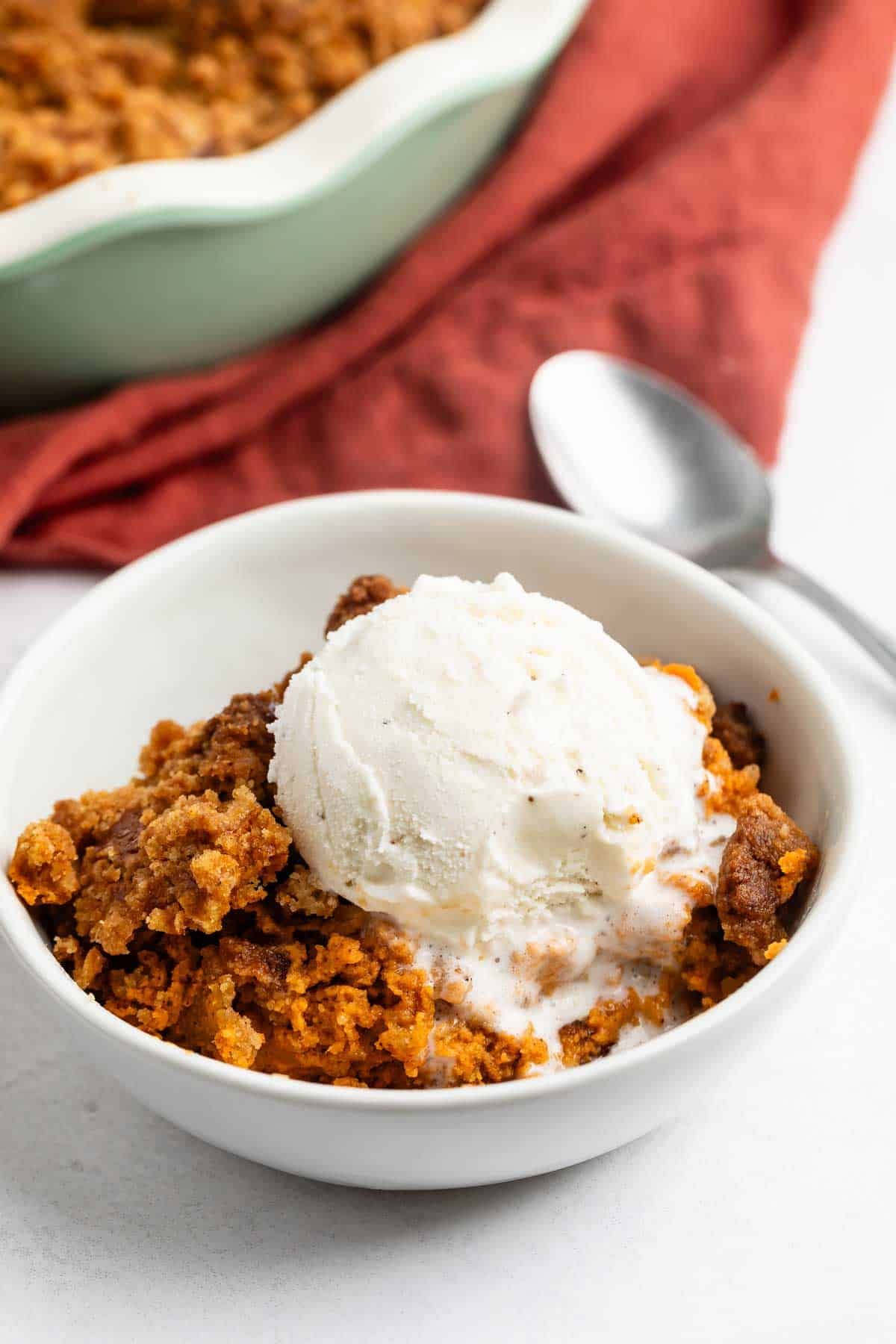 A bowl of pumpkin dump cake topped with a scoop of vanilla ice cream, with a spoon and a red cloth napkin in the background.