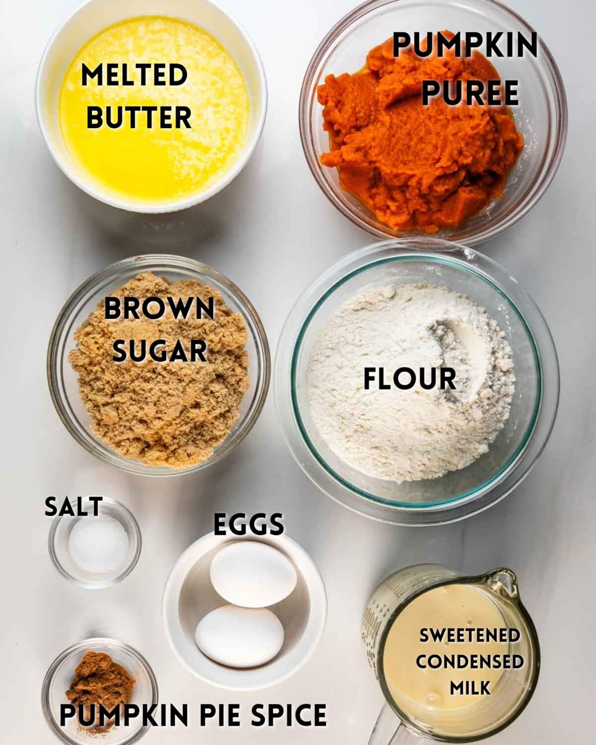Bowls of melted butter, pumpkin puree, brown sugar, flour, salt, pumpkin pie spice, two eggs, and sweetened condensed milk are arranged on a white surface, each labeled with its ingredient name.