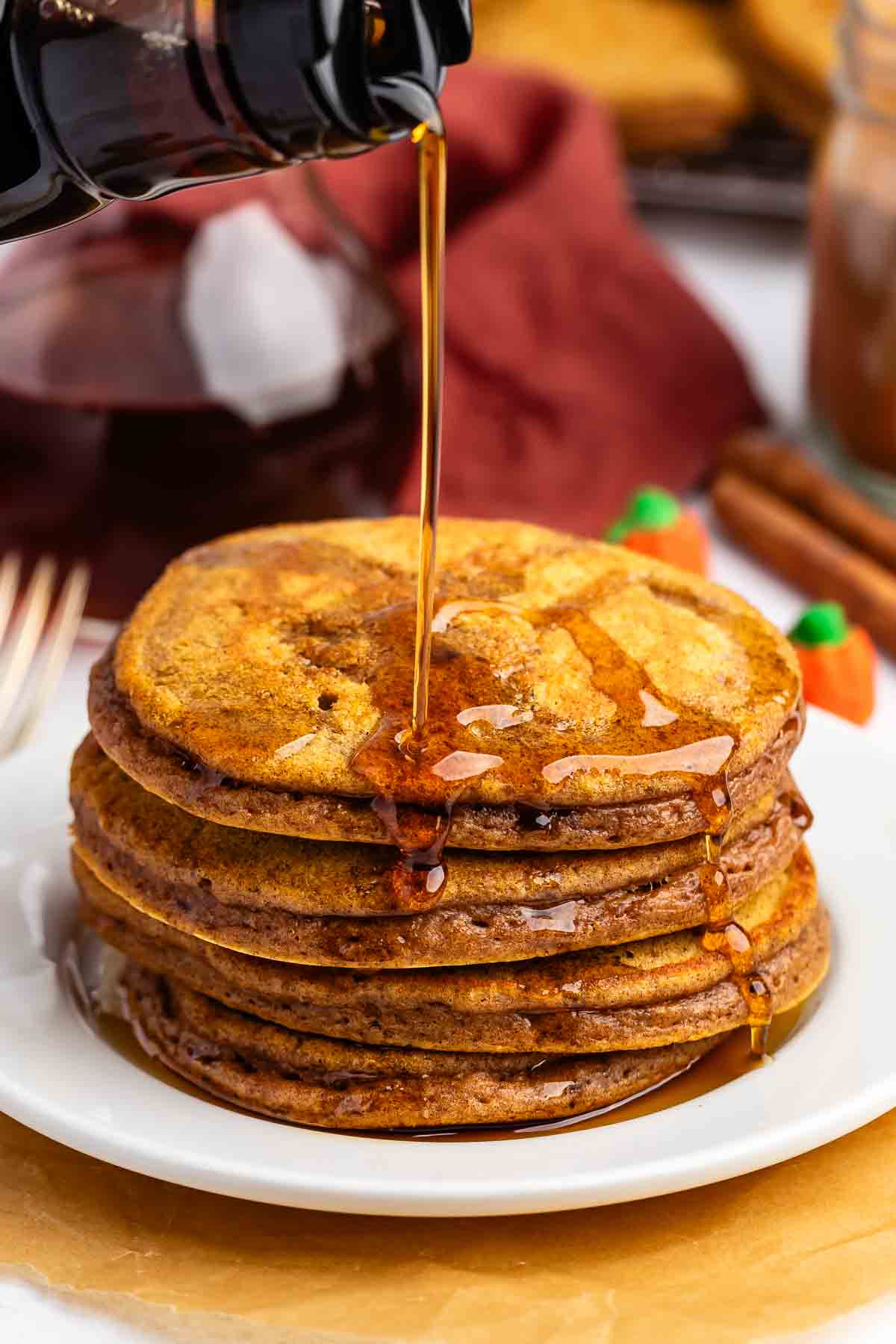A stack of pumpkin pancakes on a white plate is being drizzled with syrup.
