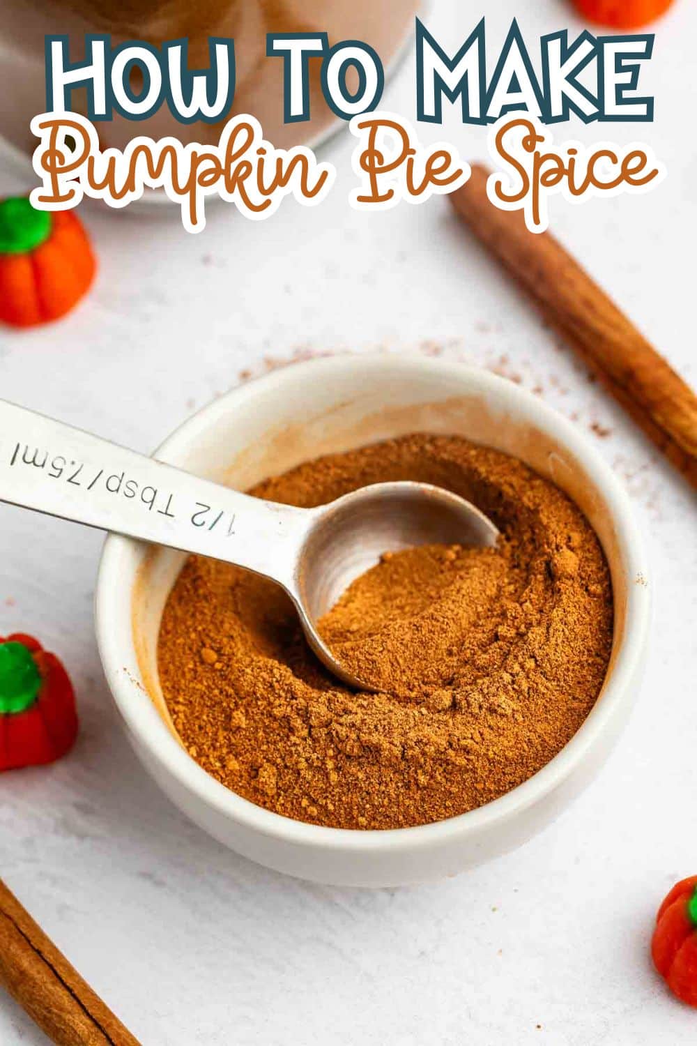 A white bowl filled with pumpkin pie spice and a metal measuring spoon, surrounded by cinnamon sticks and decorative pumpkin candies. Text at the top reads, How to Make Pumpkin Pie Spice.