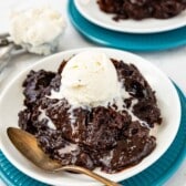 A bowl of rich chocolate cobbler, reminiscent of crockpot lava cake, is topped with a scoop of vanilla ice cream on a white plate with a vintage spoon, with another serving and extra ice cream in the background.